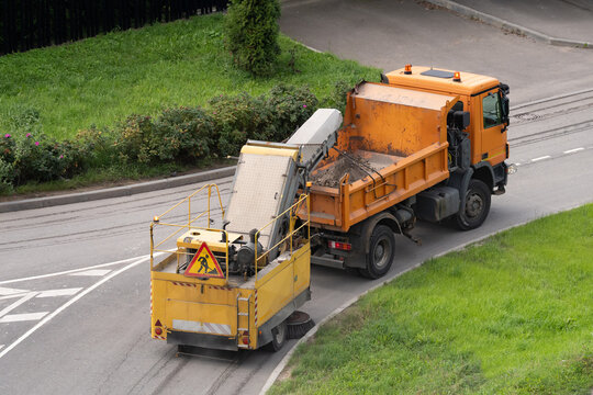 Urban road maintenance and public cleaning services in city, street sweeper machine sweeping asphalt road, cleaning debris from surface and transfers by conveyor belt directly into open dump truck.