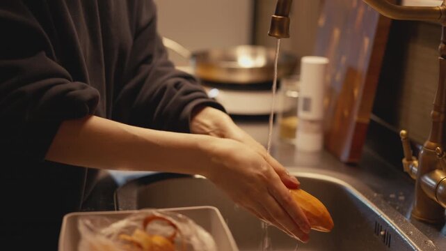 Woman washing peeled sweet potato under running water tap in kitchen sink and shaking off excess drops. Close-up of food prep process with brass faucet and stainless steel countertop in home kitchen.