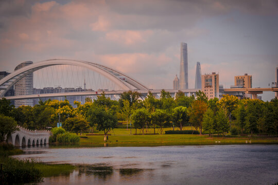 View of Expo Park with its white arched bridge over a calm lake, surrounded by lush trees and the modern Shanghai skyline under a soft sunset sky in Shanghai, Shanghai, China.