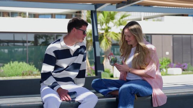 A young White couple sits face to face, shares a takeaway meal and enjoys a relaxed conversation on a modern outdoor terrace.