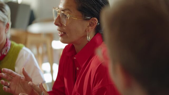 Colleague watches as woman presents ideas during a team meeting in a cafe workspace, showing business collaboration, coworking discussion, and startup planning with the group.