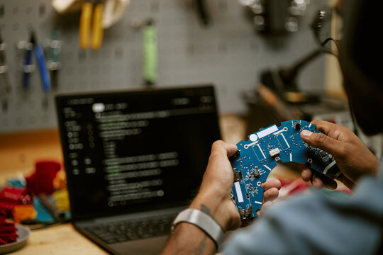 Unrecognizable engineer holding circuit board prototype while writing software code on laptop. Visual ideal for hardware engineering and tech education campaigns
