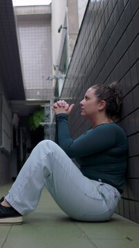 Exhausted young female resting spine against corridor wall with folded fingers finding serene stillness emotional decompression and quiet recovery after prayer