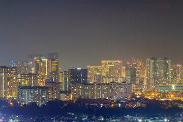 Fototapeta premium A panoramic nighttime cityscape shows dense high-rise buildings, glowing windows, rooftops, and street lights beneath a hazy dark sky with ample copy space.