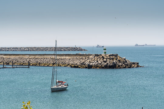 View of a sailboat anchored in the harbor near the breakwater and lighthouse in Sines, Setubal, Portugal.