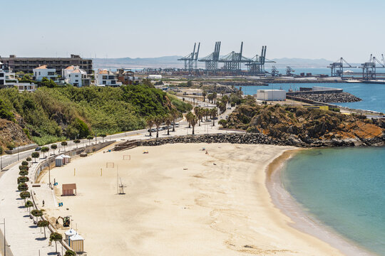 View of the sandy beach in Sines with palm trees along the promenade, rocky cliffs, and the industrial port infrastructure in the background in Sines, Setubal, Portugal.