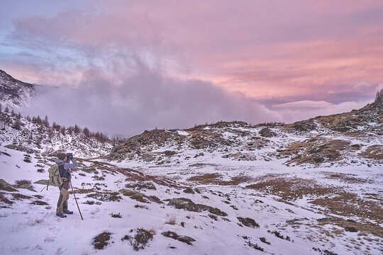 View of a hiker with a backpack and walking stick observing the snowy Vallespluga mountain landscape under a dramatic pink sunset sky in Lombardia, Italy.