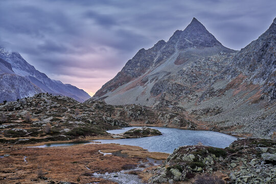 View of Engadin mountain landscape with a serene alpine lake, rocky slopes, and a dramatic sunset sky in Grisons, Switzerland.