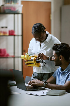 Vertical shot of African American young woman and Indian man programming robot prototype with laptop in laboratory, ideal for STEM education marketing