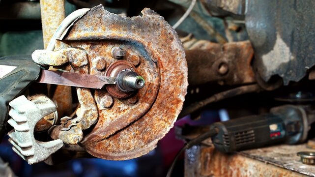View of a mechanic using a tool on a heavily rusted car brake rotor and hub assembly in a workshop in Latvia.