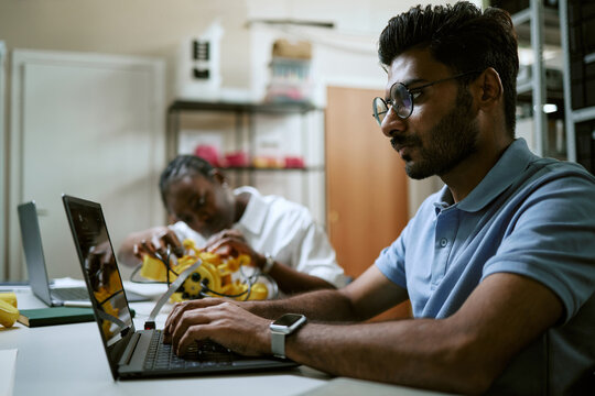 Young Indian man programming robot on laptop while African American woman student builds robotic device in tech lab, suitable for engineering education and AI research marketing