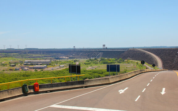 View of the massive Itaipu Dam hydroelectric structure seen from a road with surrounding green vegetation under a clear blue sky in Itaipulandia, Parana, Brazil.