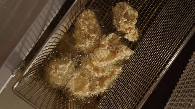 Breaded food pieces cook in a fryer basket surrounded by bubbling hot oil in a professional kitchen.