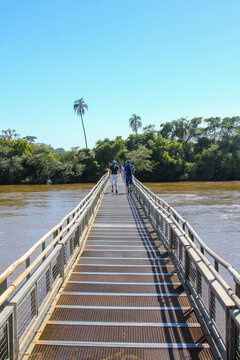 View of a metal footbridge over the Iguazu River with people walking towards the lush vegetation near Iguazu Falls in Foz do Iguacu, Parana, Brazil.