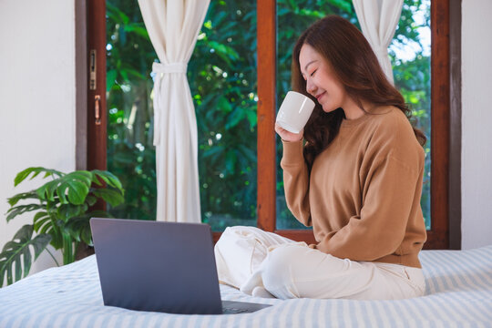 Closeup image of a woman working on laptop computer and drinking coffee while sitting on a bed at home