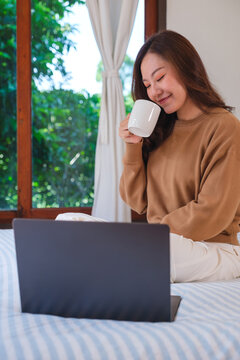 Vertical closeup image of a woman working on laptop computer and drinking coffee while sitting on a bed at home