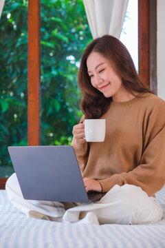 Vertical closeup image of a woman working on laptop computer and drinking coffee while sitting on a bed at home