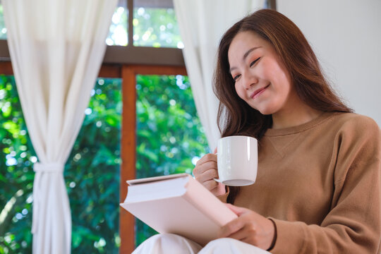 Portrait image of a woman drinking coffee and reading a book on the bed at home