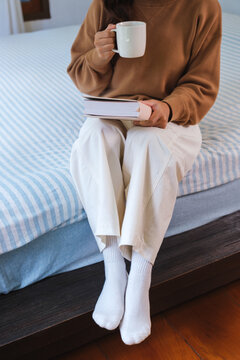 Vertical closeup image of a woman drinking coffee and reading a book on the bed at home