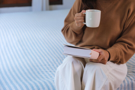 Closeup image of a woman drinking coffee and reading a book on the bed at home
