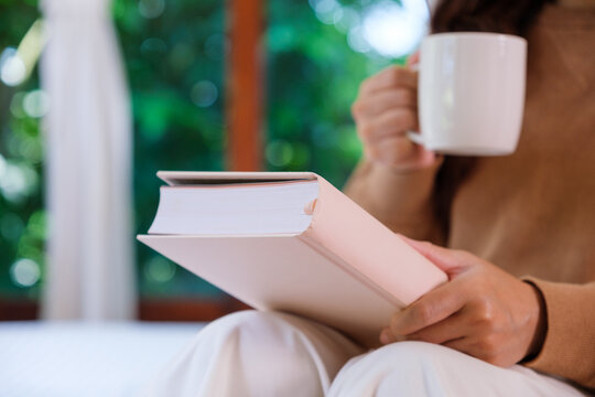 Closeup image of a woman drinking coffee and reading a book on the bed at home