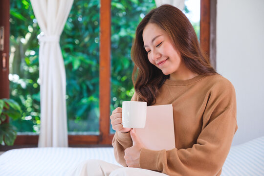 Portrait image of a woman drinking coffee and holding a book on the bed at home