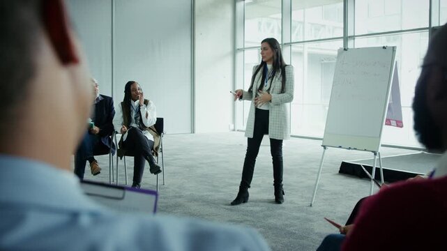 A businesswoman confidently addresses a group of attendees during a corporate meeting. They are engaged in learning and collaboration at a professional event.