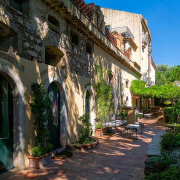 A composition highlighting terracotta floor tiles, dappled sunlight shadows, stone arch masonry, green foliage, and plaster textures in Taormina.