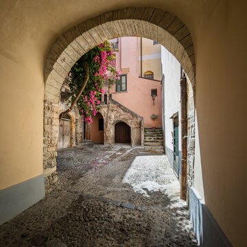 View through a historic stone archway into Cortile Mandralisca with blooming bougainvillea in Cefal&ugrave;, Sicily, Italy.