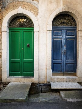 Vibrant green and blue arched wooden doors side-by-side in historic Sicily, featuring ornate iron fanlights and weathered stone steps.