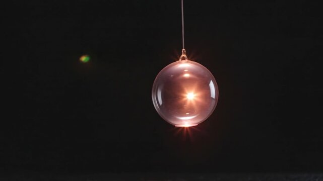 Cinematic video of a transparent hanging glass sphere illuminated by warm light with reflections against a deep black background.