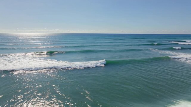 Cinematic video of gentle ocean waves rolling toward shore beneath a bright blue sky with sunlight shimmering across the water.