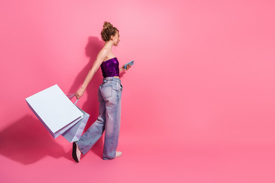 Fashionable stylish woman wearing glamorous outfit holding shopping bags and smartphone against pink background in casual pose