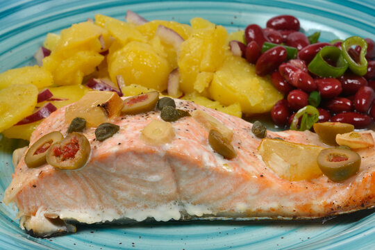 Baked salmon fillet topped with olives capers and lemon served with potato salad and red beans on a blue plate.