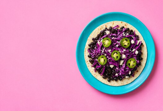 A vibrant tostada topped with black beans, purple cabbage, jalape&ntilde;o slices, and crumbled queso fresco on a blue plate against a pink background