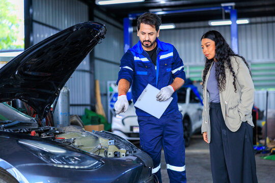 Male auto mechanic in a blue uniform holding a clipboard and explaining the car repair estimate to a female customer next to a car with an open hood in an auto repair shop.