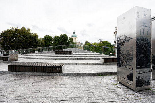 Modern stepped plaza with benches and reflective monuments in Radom, Poland, framed by trees and a historic church dome in an urban public square