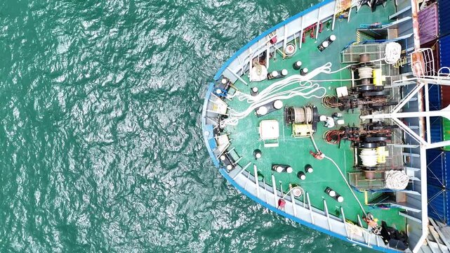 multiple ship crew members in bright safety orange working with massive ropes and heavy machinery on the vibrant green deck of a large vessel's bow, with blue water surrounding