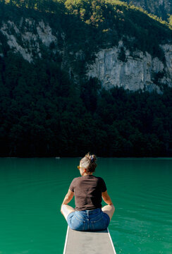 Back view of a young woman sitting cross-legged at the edge of a diving board overlooking a mountain lake