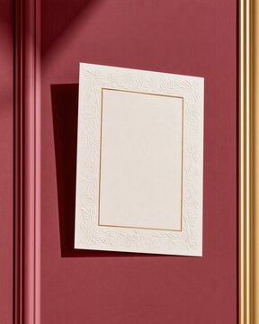 White embossed invitation card with gold border on deep red textured wall, soft shadow and golden frame edge visible