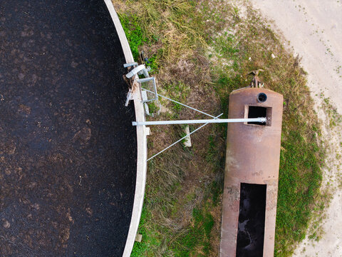 Aerial view of a large circular industrial tank filled with dark liquid next to a rusted metal container in a rural setting in Virpe, Ventspils novads, Latvia.