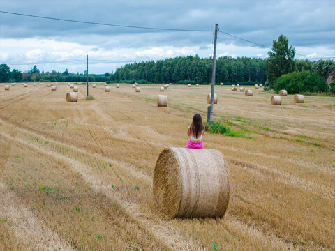 Aerial view of a woman sitting on a large hay bale in a vast harvested field under a cloudy sky in Latvia.