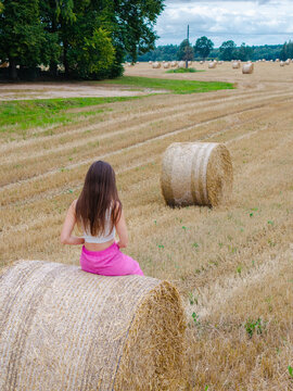 Aerial view of a woman sitting on a hay bale in a harvested field with scattered hay bales and trees in the background in Latvia.