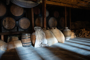 Dark cargo hold of a wooden vessel filled with vintage wine barrels, burlap sacks, and ceramic jugs. © Adolf