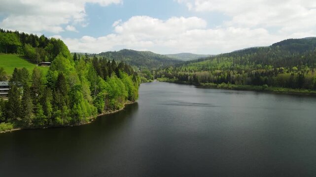 Aerial drone view showing a tranquil lake surrounded by lush green wooded hills. Rolling terrain stretches across the horizon beneath a partly cloudy sky in Wigry Poland
