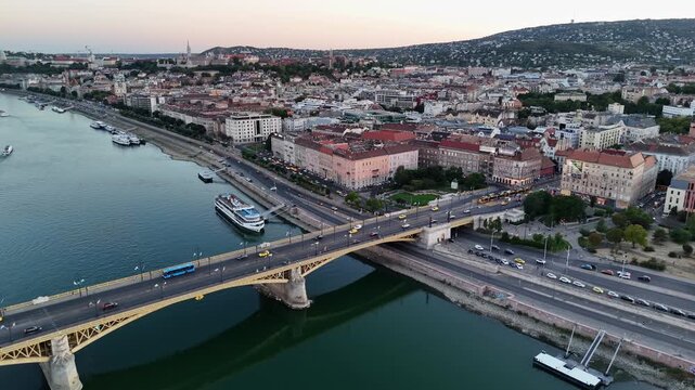 Budapest, Hungary, Margaret Bridge over the Danube with yellow tram, traffic and boats along the lower quay at sunset, framed by Buda Hills in a summer drone view.