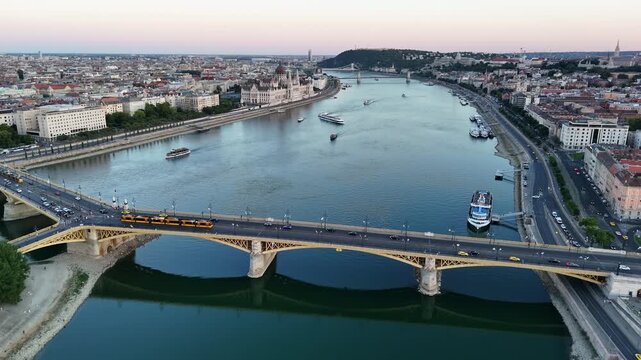 Budapest, Hungary, with the Hungarian Parliament and Margaret Bridge along the Danube at dusk, city skyline, boats, tram traffic and waterfront streets seen from a drone.