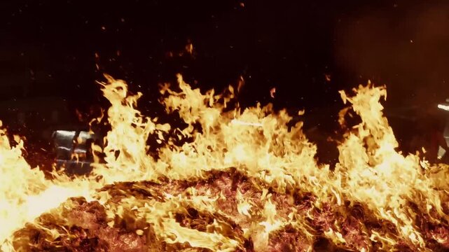 Close-up static shot of bright, chaotic flames rolling across a dense bed of burning paper offerings and glowing embers during a nighttime ritual.