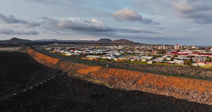 Wide drone view of Grindavik, Iceland, showing a long orange lava defense berm and road separating the residential town from a vast black lava field beneath cloudy evening skies.