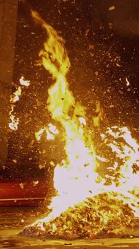 Vertical close-up static shot of a powerful flame vortex spiraling upward from a street bonfire, surrounded by a dense cloud of swirling burning joss paper and bright sparks against the night.